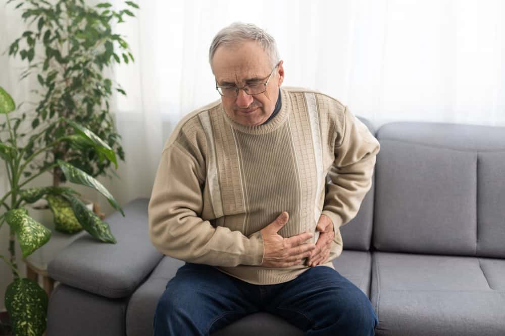 An older man sitting on a gray sofa in FL holds his stomach with a pained expression, suggesting abdominal discomfort. A green houseplant is visible beside him, highlighting the need for primary care Tallahassee residents can trust.