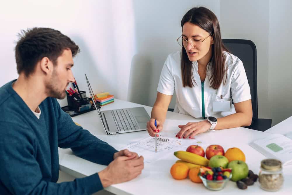A nutritionist in a white coat discusses a meal plan with a man at a desk in FL, with assorted fruits, nuts, a laptop, and documents—highlighting the importance of nutrition in primary care Tallahassee settings.