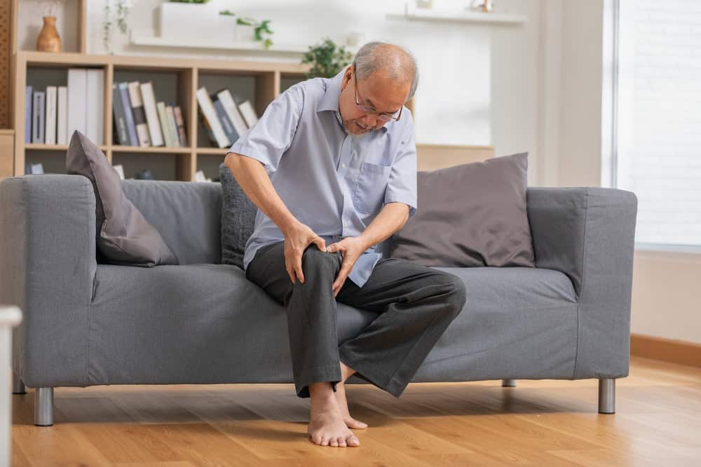 An elderly man sits on a gray sofa in a bright, modern room, holding his knee with a pained expression—suggesting he may benefit from primary care in Tallahassee, FL. He is barefoot, with a bookshelf visible in the background.
