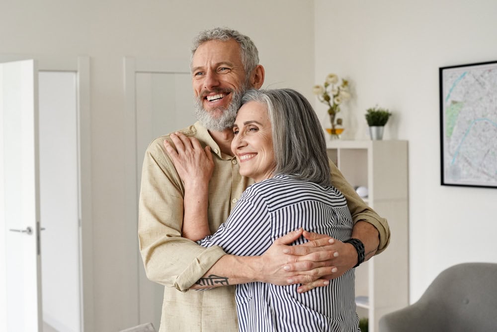 An older couple with gray hair smiling and hugging each other in a bright, modern FL living room. They appear happy and relaxed, sharing a warm, affectionate moment together—enjoying life with excellent primary care Tallahassee offers.