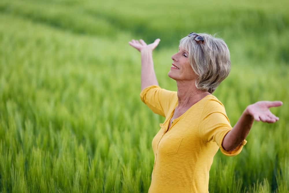 A smiling older woman with short gray hair stands in a lush green field, wearing a yellow shirt and raising her arms joyfully while looking upward, embracing the vitality supported by quality primary care in Tallahassee, FL.
