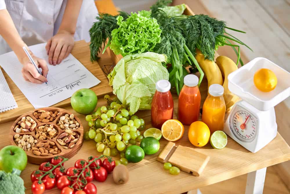 A person writes on a clipboard near a table filled with fresh vegetables, fruits, nuts, bottled juices, and a kitchen scale with a lemon on it—suggesting meal planning or nutrition tracking for primary care Tallahassee in FL.