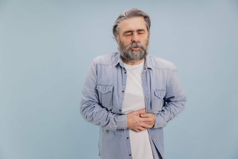 A middle-aged man with gray hair and beard stands against a light blue background, eyes closed and holding his stomach with both hands, appearing to be in pain—highlighting the need for primary care in Tallahassee, FL.