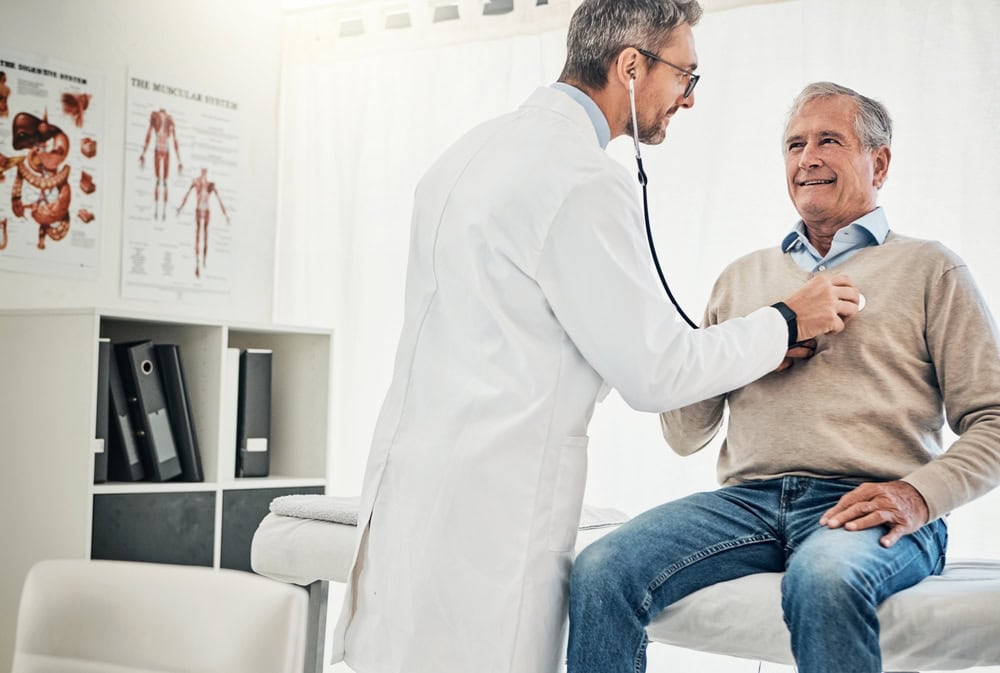 A primary care doctor in FL uses a stethoscope to examine an older man sitting on an exam table in a Tallahassee medical office. The man smiles as his chest is listened to, with medical posters and shelves visible in the background.