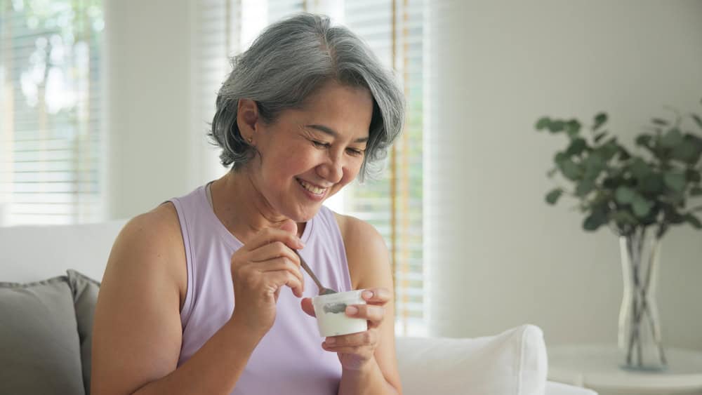 Smiling older woman with short gray hair sits on a sofa, enjoying a cup of yogurt in her bright, cozy living room. Soft natural light and lush plants fill the space—reflecting the comfort of primary care Tallahassee, FL, offers.