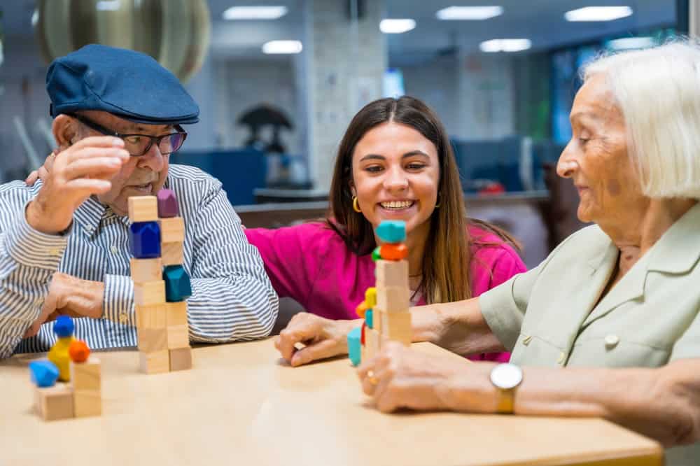 A young woman smiles while sitting at a table with two older adults who are stacking colorful blocks, engaging in a playful activity together in a bright, indoor primary care Tallahassee setting.