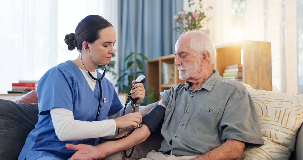 A nurse in blue scrubs checks the blood pressure of an elderly man sitting on a couch at home. Providing primary care in Tallahassee, FL, the nurse uses a stethoscope and cuff while the man looks down at his arm.