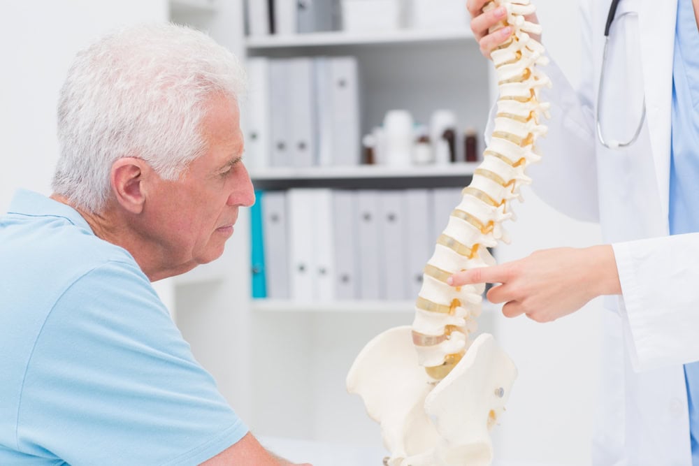 An older man listens as a doctor in a lab coat explains using a model of a human spine in a primary care Tallahassee, FL medical office, with shelves and binders in the background.