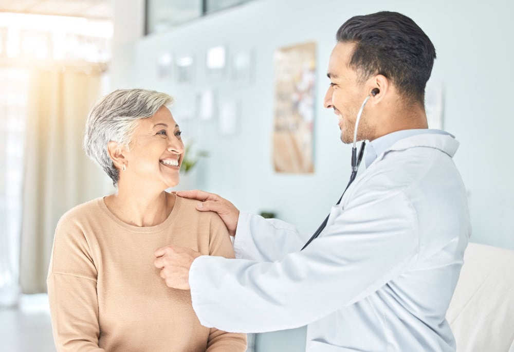 A smiling doctor with a stethoscope examines an older woman in a bright medical office, reflecting the friendly, patient-focused atmosphere of primary care in Tallahassee, FL.