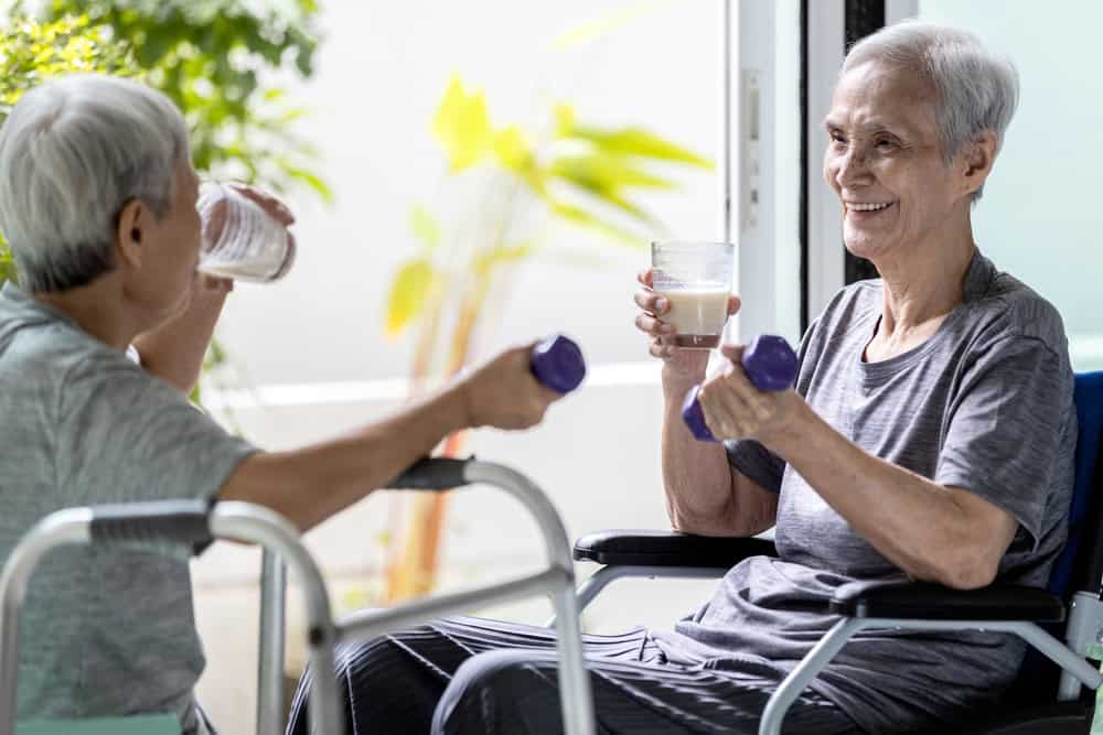 Two elderly women sit facing each other indoors, smiling. One holds a glass of water and a walker; the other, in a wheelchair, lifts a small dumbbell—reflecting exercise, healthy habits, and support available through primary care in Tallahassee, FL.