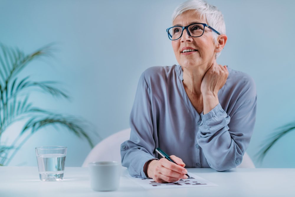 An older woman with short gray hair and glasses sits at a desk in primary care Tallahassee, FL, smiling thoughtfully while holding a pen over a piece of paper. A glass of water, coffee cup, and green plants are in the background.