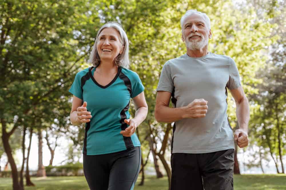 A smiling older woman and man are jogging together outdoors in a sunny Tallahassee, FL park, surrounded by green trees. Both wear athletic clothing and appear happy and healthy—enjoying the benefits of primary care in Tallahassee, FL.