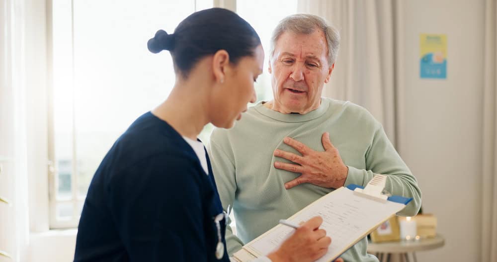 A healthcare professional writes on a clipboard while an older man, appearing concerned, holds his chest and speaks in a bright FL primary care Tallahassee clinic.
