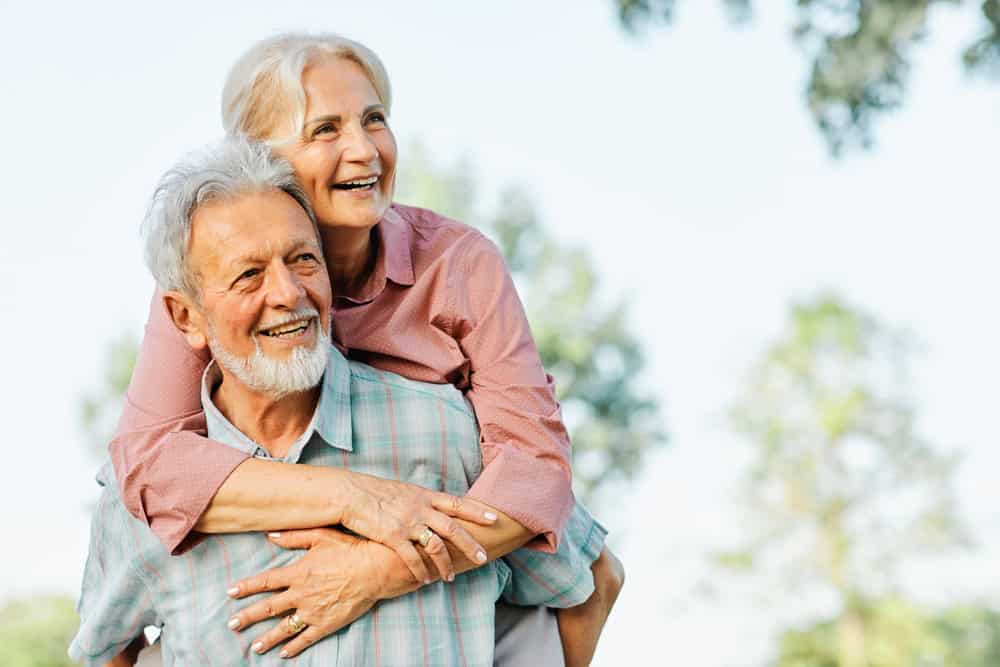 An older woman smiles while hugging an older man from behind outdoors in FL. Both appear happy, enjoying the trees and blue sky—perfectly capturing health and joy near primary care Tallahassee.