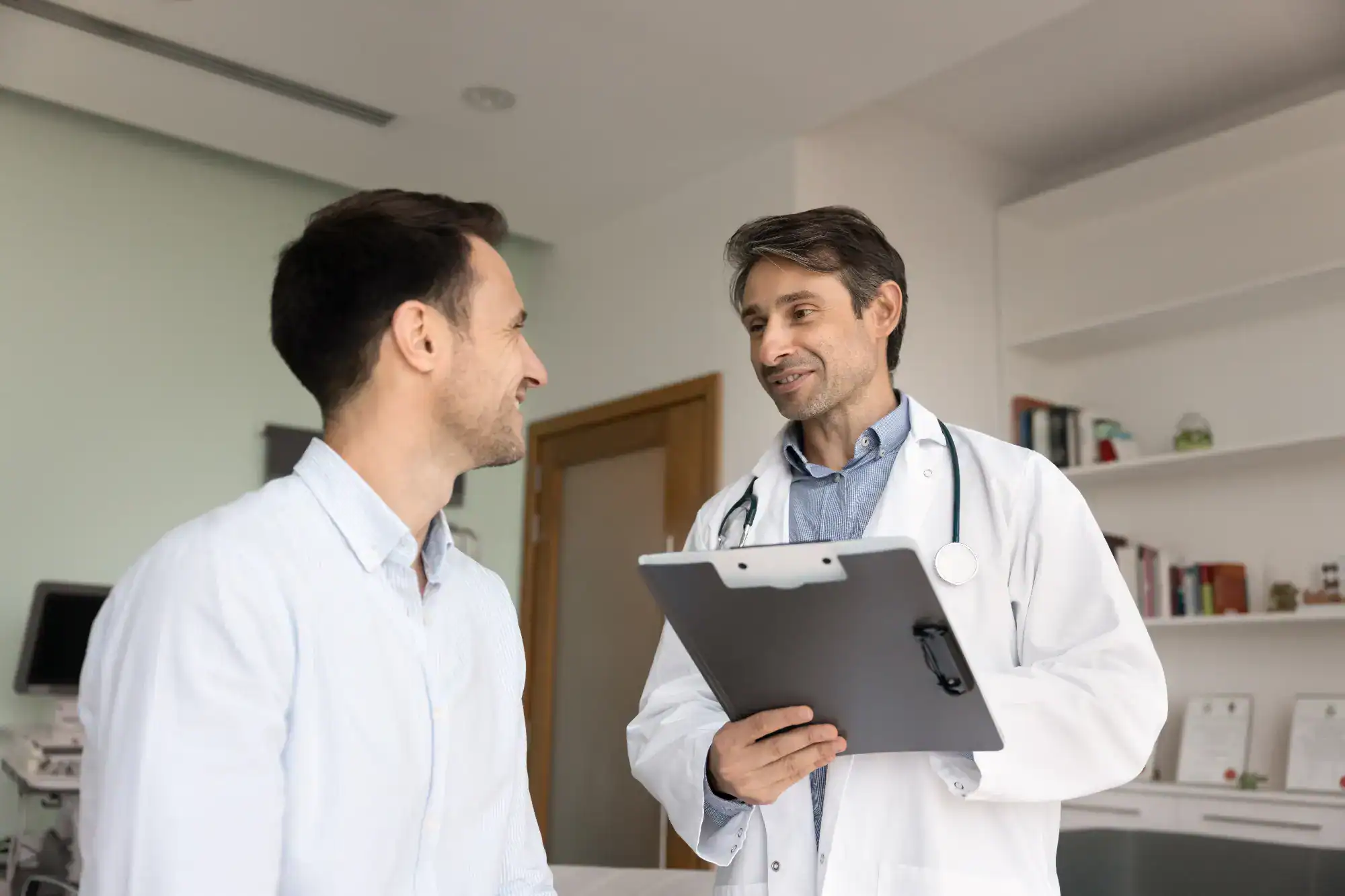 A primary care doctor in Tallahassee, FL, wearing a white coat, holds a clipboard and smiles while talking to a male patient in a medical office. Both appear engaged in a friendly conversation.