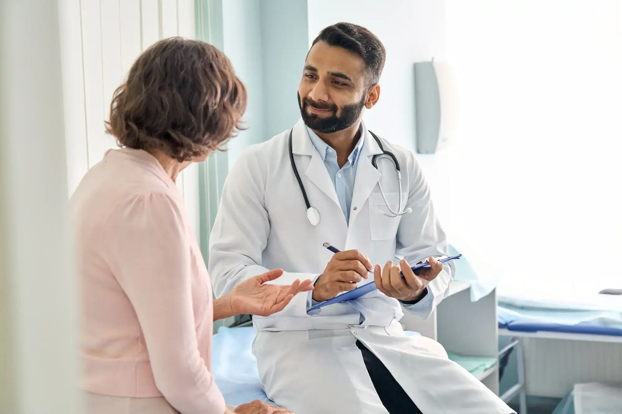 A FL primary care doctor with a stethoscope and clipboard sits and smiles while talking to an older female patient in a Tallahassee medical office. The patient gestures with her hand as they discuss her health.