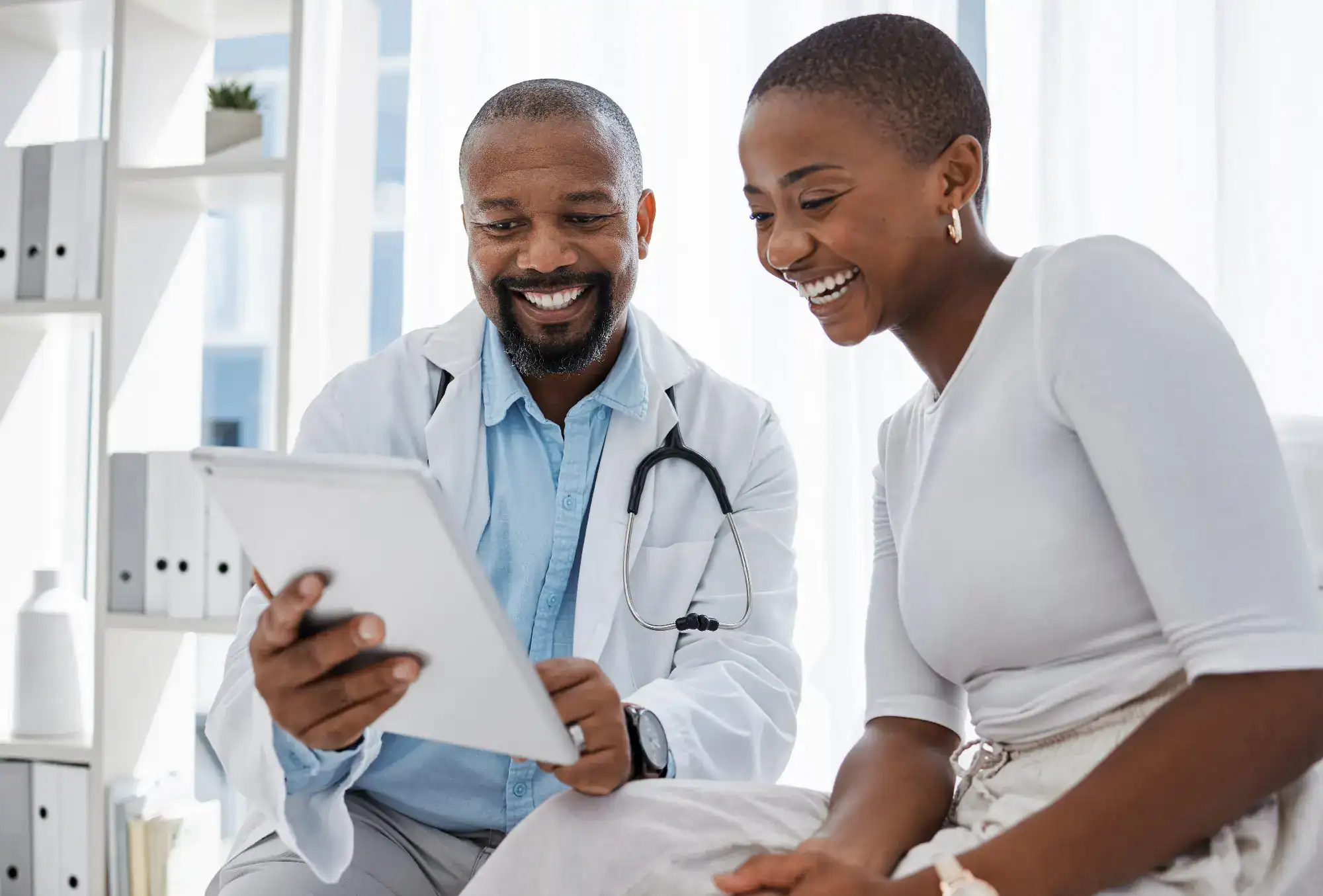 A smiling doctor shows a digital tablet to a happy patient during a consultation in a bright medical office, reflecting the positive and supportive atmosphere of primary care Tallahassee, FL. Both appear engaged and comfortable.