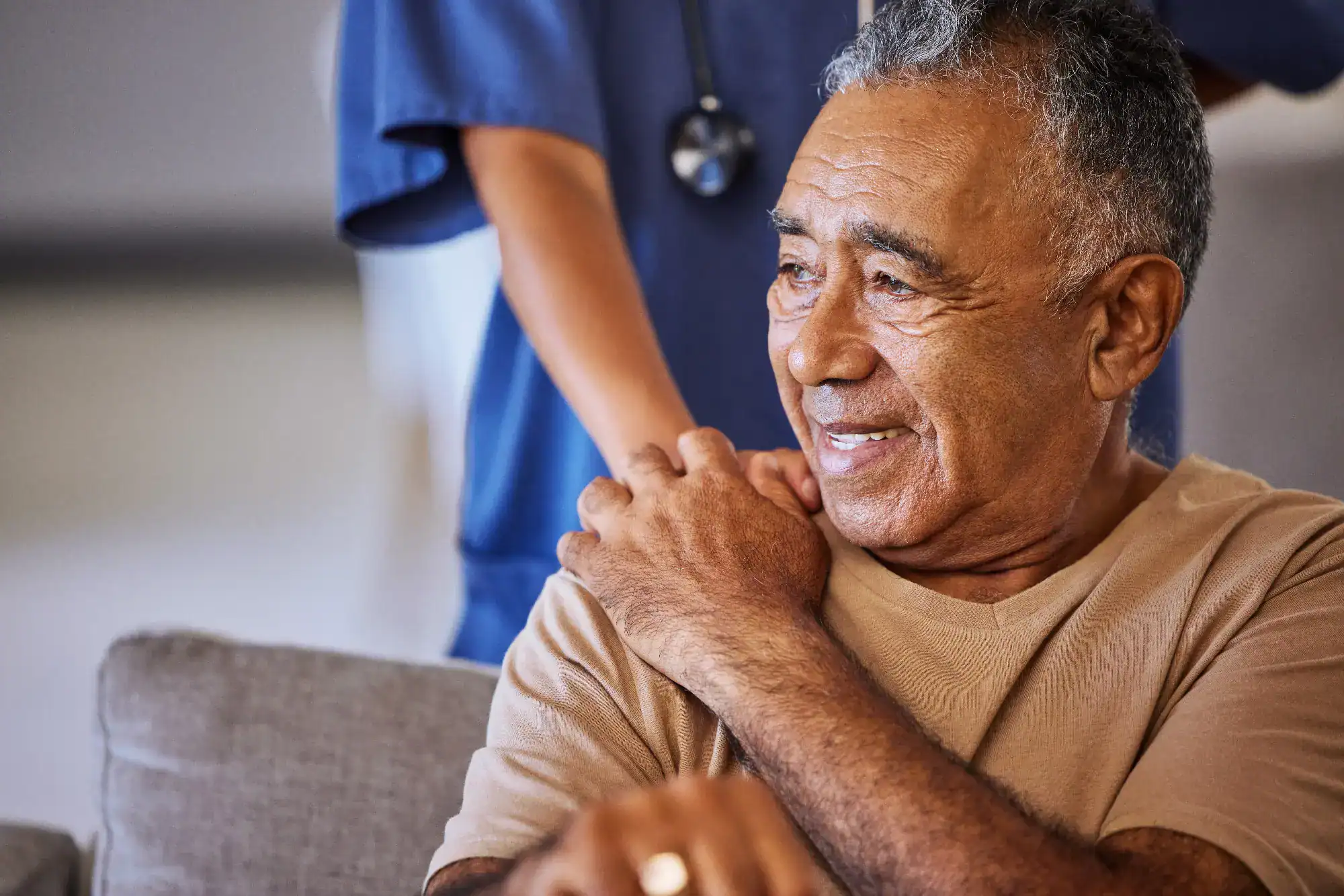 An older man smiles while sitting on a couch, as a nurse in blue scrubs from FL primary care Tallahassee gently rests a hand on his shoulder, offering comfort and support.