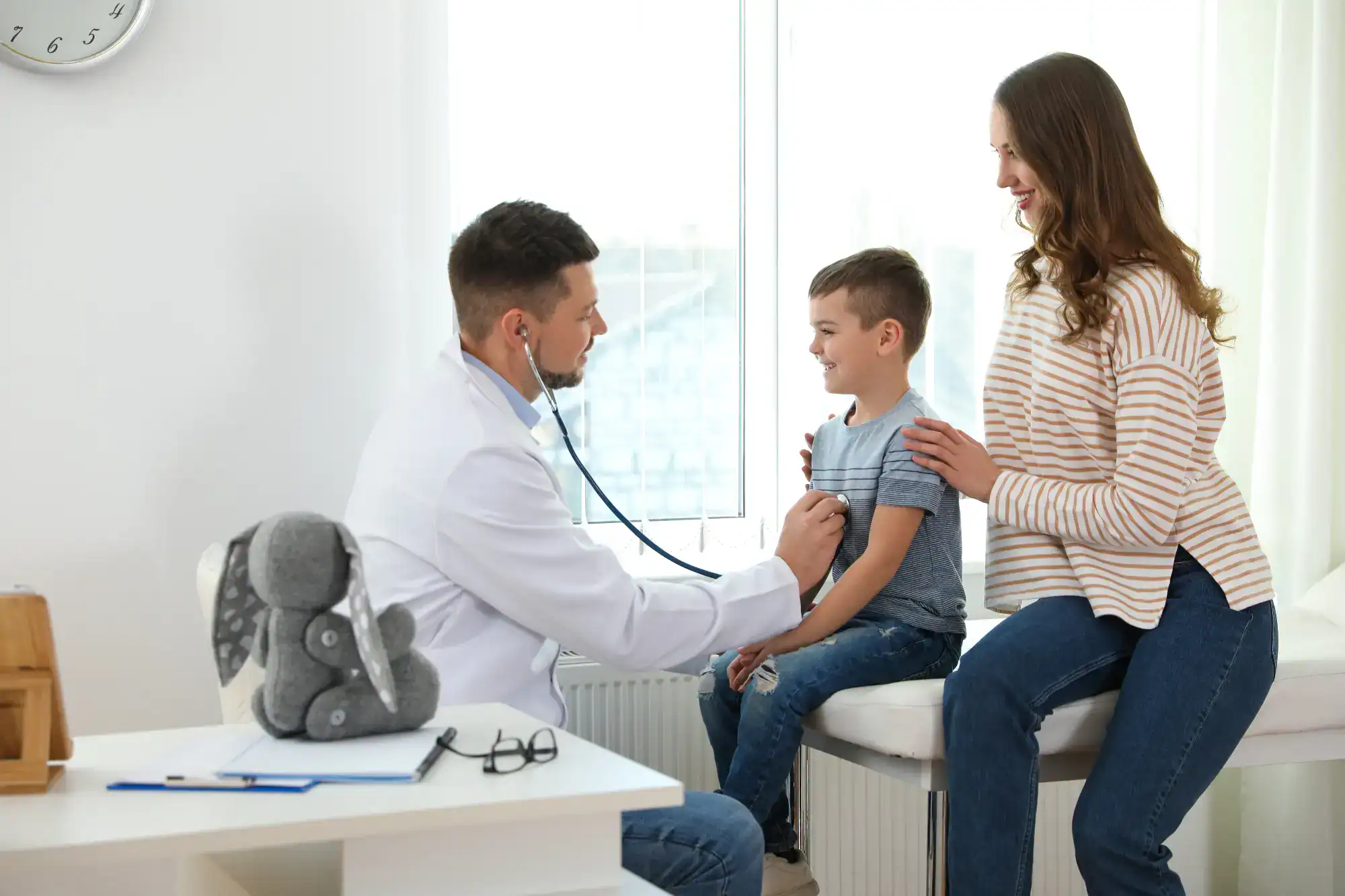 A doctor uses a stethoscope to check a smiling young boy in a bright FL primary care Tallahassee office, while his mother sits nearby. A plush toy rests on the doctor's desk, adding comfort to the visit.