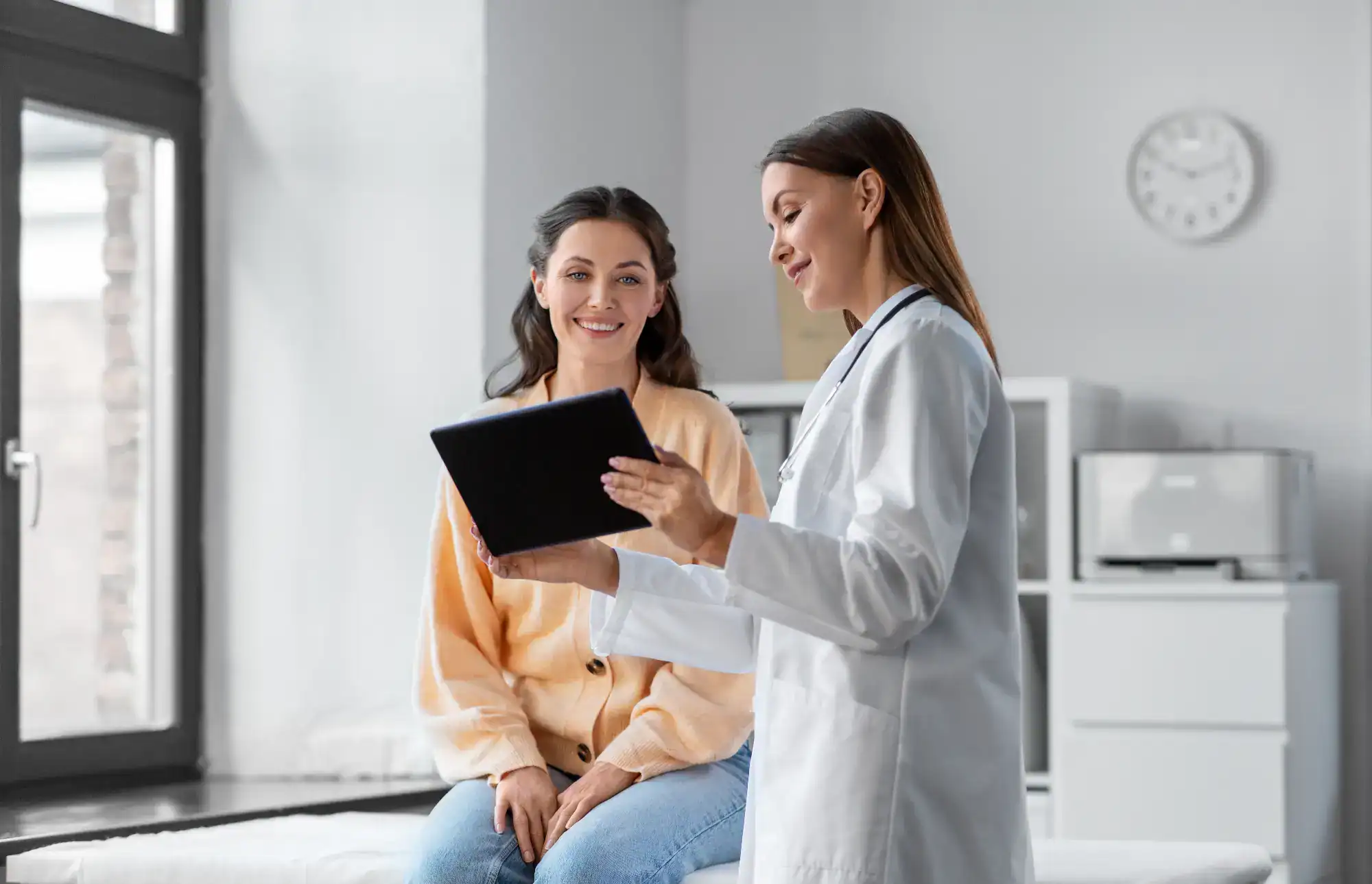 A doctor in a white coat shows information on a tablet to a smiling woman sitting on an exam table in a bright FL primary care Tallahassee medical office.