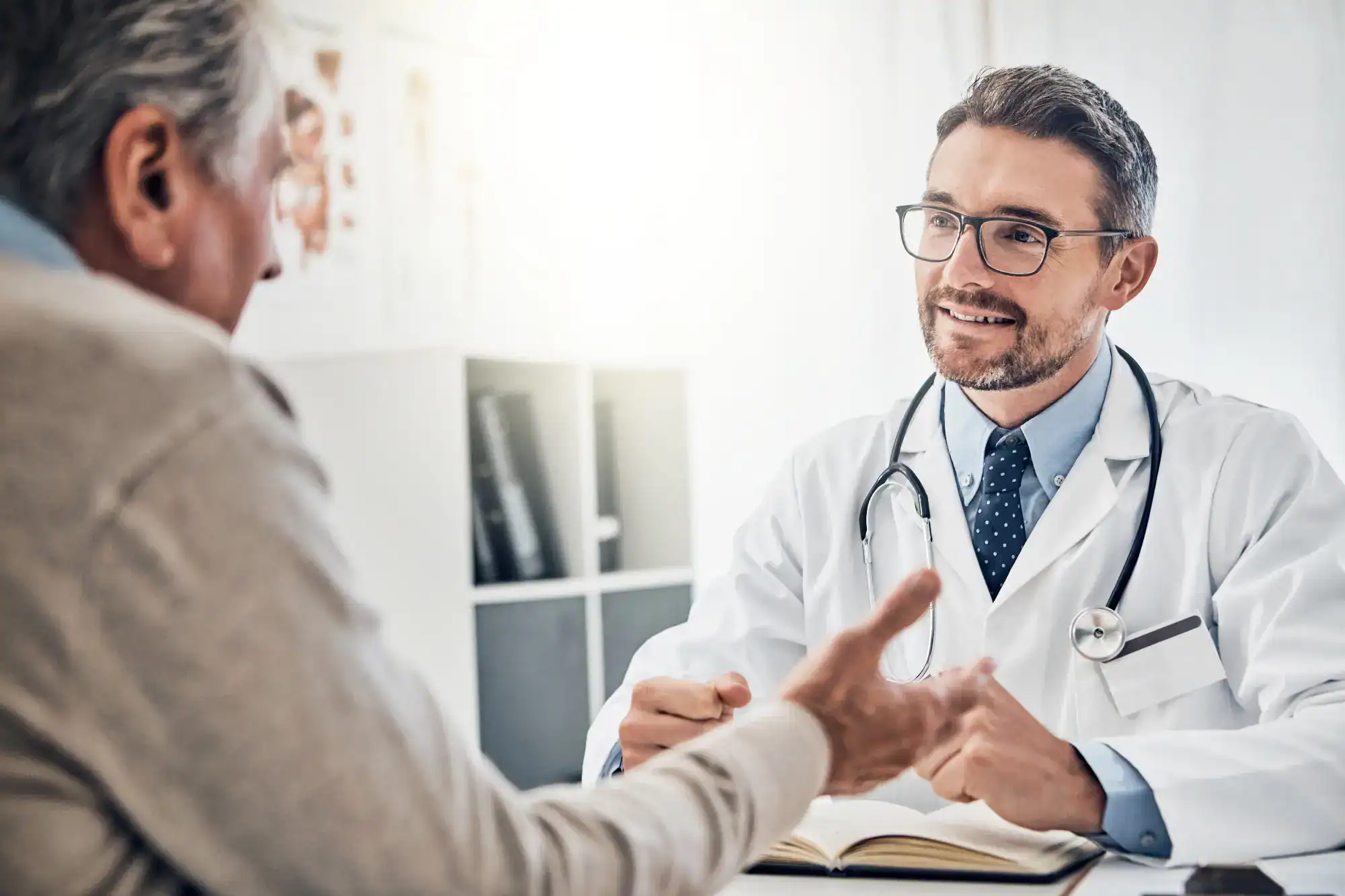 A male doctor in a white coat and stethoscope listens and smiles while consulting with an older male patient, who gestures during a discussion in a bright primary care Tallahassee, FL medical office.